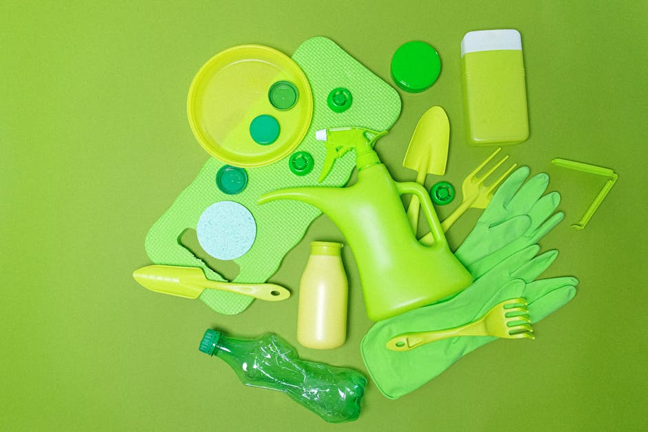 A collection of garden maintenance tools and supplies in various shades of green arranged on a green background. Included are a watering can, a pair of rubber gloves, a small trowel, a rake, a spray b