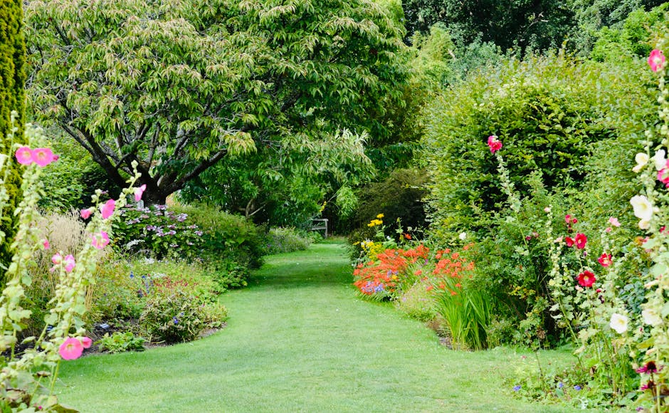 A lush garden scene featuring a well-maintained, vibrant green lawn bordered by a variety of colorful flowering plants and shrubs. On the left side, tall pink hollyhocks and other flowering perennials